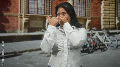 Obraz na płótnie Woman covers mouth with hand, standing by parked bicycles in front of a brick building on a street; surprise intimacy