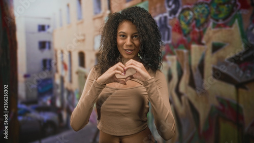 Teenage woman makes a heart with her hands and smiles  exposed midriff visible  standing by graffiti covered wall on a street  joyful self love.