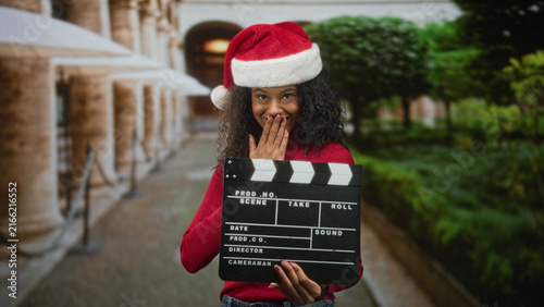 Obraz na płótnie Teenage african american woman with curly hair and santa hat holds a clapboard and covers mouth with hand in studio; holiday surprise