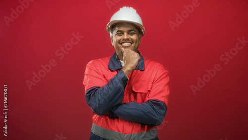 Hispanic construction worker in hard hat and red safety uniform smiling with hand on chin in studio  confidence professionalism.