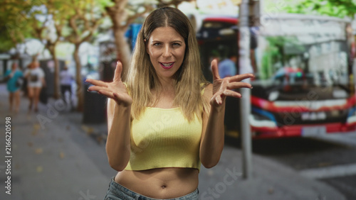 Konstfotografi Woman with yellow tanktop finger to lips, midriff visible and gaze forward, standing by a busstop on a street; flirtatious confidence