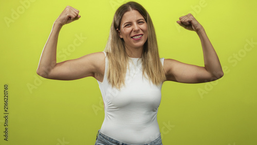 Cuadro en lienzo Woman flexes arm showing bicep and clenched fist in studio wearing white sleeveless top and denim jeans; confidence strength empowerment