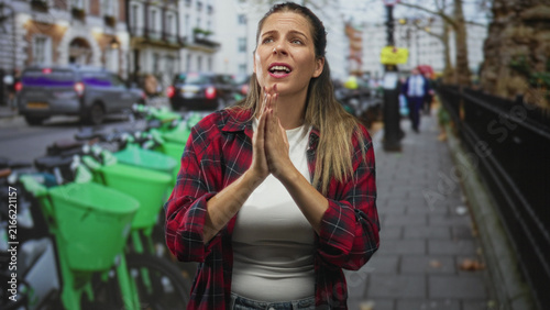фотографія Woman in red plaid shirt with hands clasped in prayer on a busy city street beside green rental bikes and passing cars, pleading expression; hope