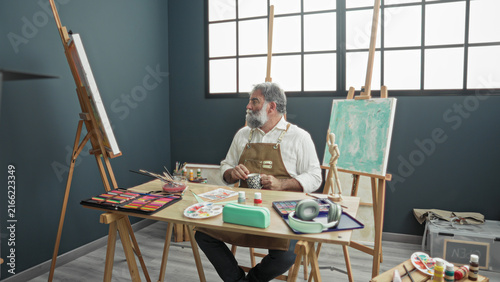Фотография Man painter with hoary beard in apron stirs mug with right hand at a paint strewn table beside easel in studio; quiet contemplation