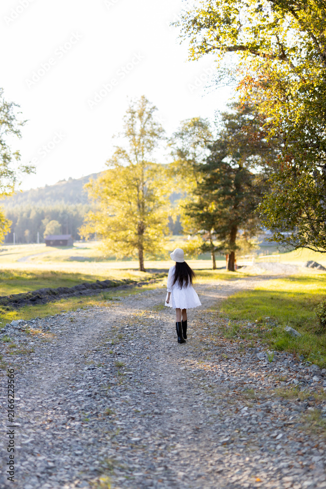 Fototapeta premium A young woman strolls down a gravel path surrounded by vibrant trees as the golden sunlight filters through leaves. The serene landscape invites peace and reflection.