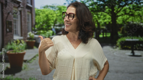 Bild auf Leinwand Woman smiling points thumb over shoulder toward building courtyard with potted plants and brick wall, wearing glasses and blouse; confidence friendly invitation