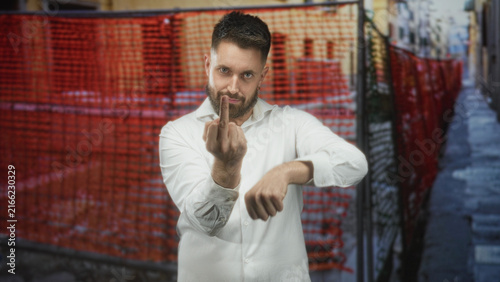 Bild auf Leinwand Man in white shirt shows middle finger and clenched fist, bare hand prominent, in narrow street with red construction fence and alley behind him; defiance