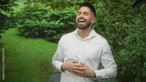 Obraz na plátne Hispanic man with beard laughing, hands on stomach, wearing white shirt on a forest path with dense green shrubs; joy and relief