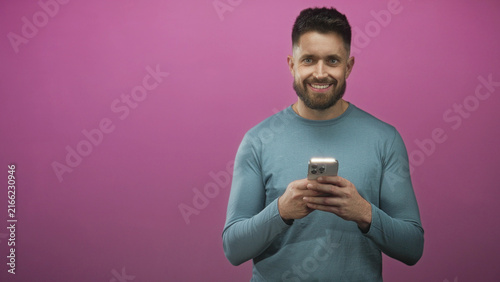 Fotografie Hispanic man with beard holds smartphone with both hands, texting and glancing up from screen in a pink studio backdrop; casual connection happy