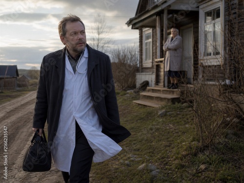 Male doctor carrying a medical bag while walking toward a rural house. Healthcare professional making a home visit in a remote community. Primary care and outreach service concept.