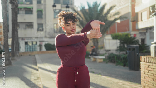 Fotografie Woman stretching hands across chest while standing on sunlit street wearing maroon activewear and sneakers; calm focus wellness