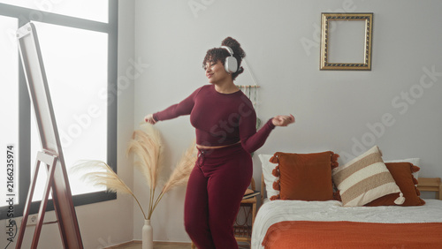 Fotografie Woman brunette dancing with headphones, pointing and swaying in a bright bedroom inside a building with bed and pillows; joyful relaxation