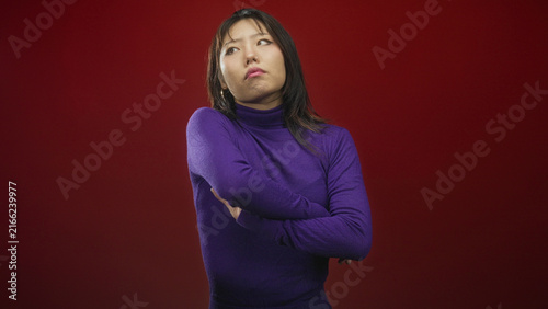 Принти на полотні Woman in purple turtleneck with arms crossed and one hand near temple, posing against a red studio backdrop; annoyed defiance