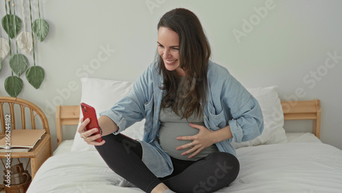 Fotografía Pregnant woman sitting crosslegged on bed holding red smartphone with one hand and other hand on belly smiling in a studio; expectant joy bonding