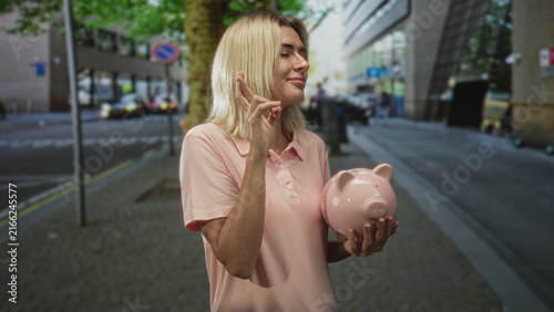 Obraz na plátně Young woman holds pink piggy bank while crossing fingers with her hand on a city street; hopeful savings future