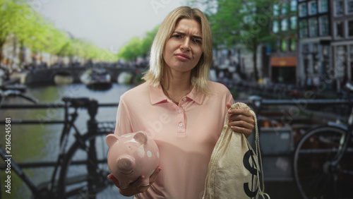 Cuadro en lienzo Woman holds piggy bank and dollar sign money sack on a street bridge, frowning as she inspects the coins; financial worry disappointment