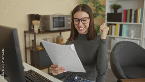 Foto Young woman holding papers, smiling and fistpump in studio workspace with computer, bookshelf and chair; success joy