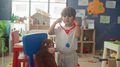 Konstfotografi Boy holds red toy stethoscope to plush monkey and lifts earpieces to his ears while standing in a playroom building; playful curiosity