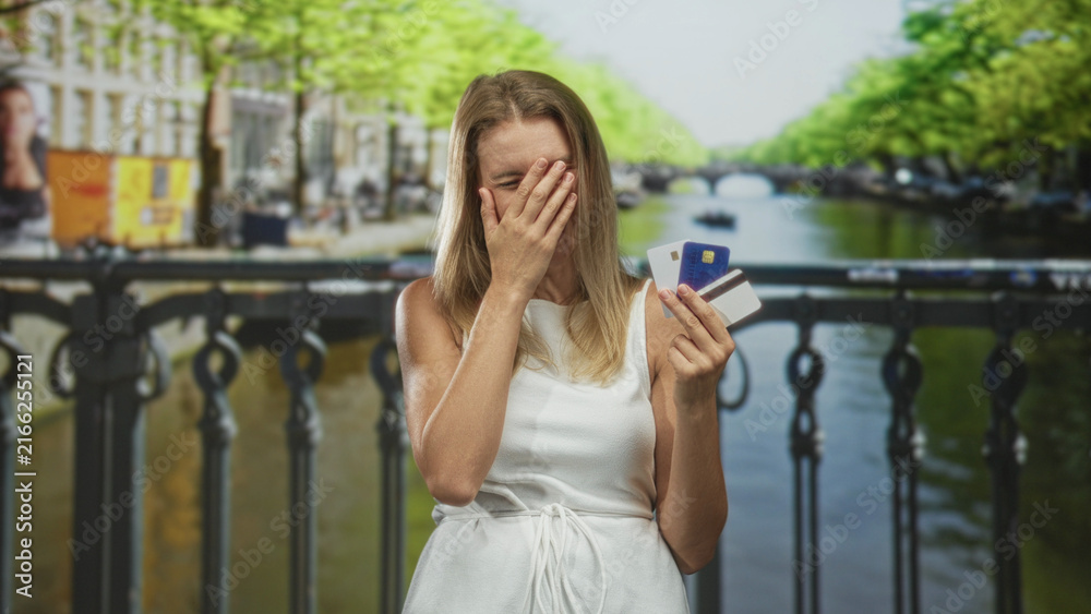 Fototapeta premium Blonde woman holding creditcard stack, hand covering face on bridge over canal; financial worry embarrassment.