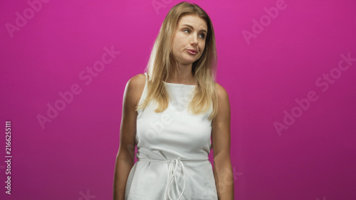 Фотография Woman plugging ears with fingers and shrugging in white dress against a pink studio backdrop; annoyance defiance