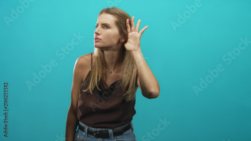 Fotografie Woman cups hand to ear listening intently in studio with turquoise backdrop, wearing brown camisole and jeans; curiosity focus