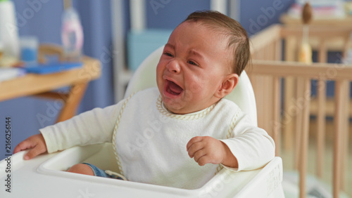 Obraz na płótnie Hispanic baby in highchair crying with open mouth and outstretched hands near crib and toys in building; upset need comfort