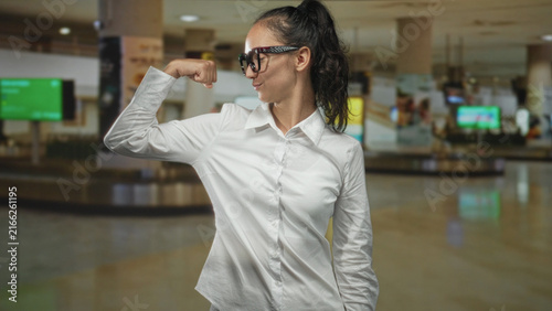 Quadro su tela Woman smiling wearing glasses flexes bicep in white shirt at airport baggage claim; confidence empowerment