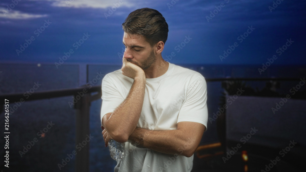 Fototapeta premium Man with beard in white shirt holding water bottle, hand on chin leaning on balcony railing of building overlooking ocean at dusk; quiet contemplation.