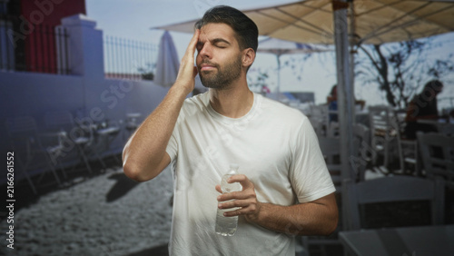 Fotografie Hispanic man with beard holding a water bottle, finger to forehead in street cafe patio; pensive reflection