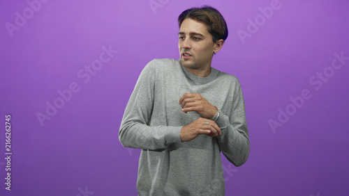 Obraz na plátne Man in studio clutching his wrist and holding a hand to chest against a purple backdrop, wearing a gray sweater and ring; unease vulnerability