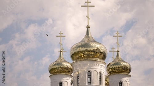 Three golden onion domes topped with orthodox crosses stand against soft blue sky with scattered white clouds. Bird in flight adds motion
