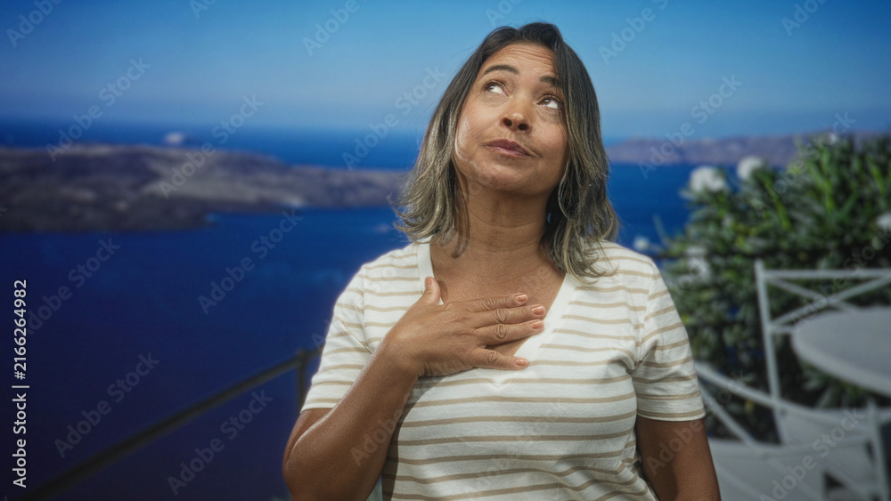 Fototapeta premium Woman middleaged hispanic with hand on chest looking up on building balcony with white railing, striped shirt and sea view; quiet gratitude.