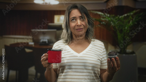 Canvastavla Woman holding red cup and chocolate muffin with eyes closed and grimace in building; hesitation disgust