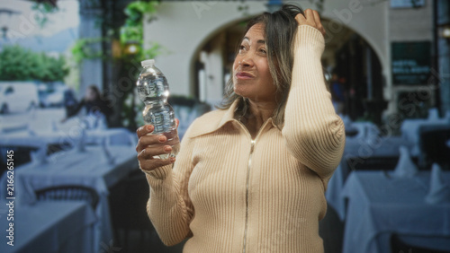 Obraz na plátně Woman holding a plastic water bottle and touching hair while squinting in a restaurant inside a building archway with tables and white linens visible; refreshment relief