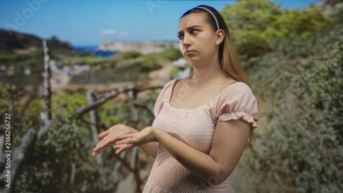 Fotografie Woman with palms up in shrug gesture on a coastal cliff trail beside a wooden railing, hands extended, wearing pink top and headband; indifference pause reflection