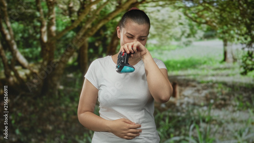 Obraz na płótnie Woman holding gamepad and pinching nose with one hand while holding stomach with the other in forest, grimacing at an unpleasant scent; disgust nausea