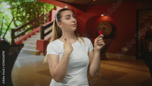 Fotografía Woman in white tshirt holding a small red heart with fingertips in a building lobby, pointing finger to chest while studying it; hesitant love