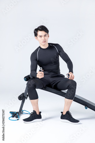 Young man with hand grippers sitting on workout bench in studio