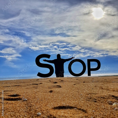 A silhouetted person stands with outstretched arms on a sandy beach under a cloudy sky, forming the word stop.