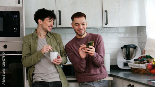 Fotografie Happy gay couple smiling and interacting with smartphone in their modern kitchen