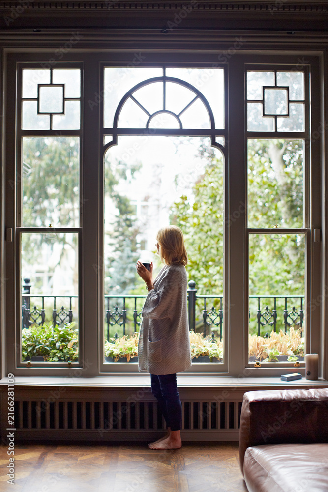 Fototapeta premium Thinking, relax or woman at window with coffee, morning reflection or calm moment on weekend break. Chill, back and person in house with mug, peaceful perspective and mindfulness at start of day.