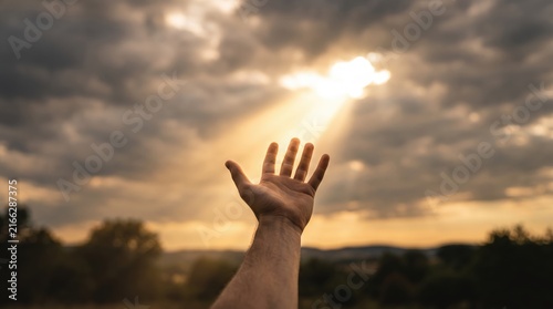Human hand reaching up towards bright sunlight breaking through dark dramatic clouds at sunset, symbolizing hope, faith, spiritual connection, and freedom, with a natural landscape