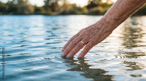 of elderly hand touching water surface creating ripples, senior skin texture, golden hour sunlight reflection on lake, peaceful nature connection, aging, retirement lifestyle