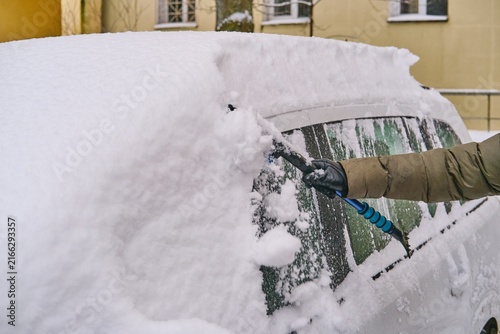 A man in a dark green jacket brushes snow off a car parked in a yard.