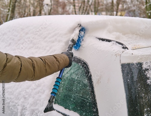 A man in a dark green jacket brushes snow off a car parked in a yard.