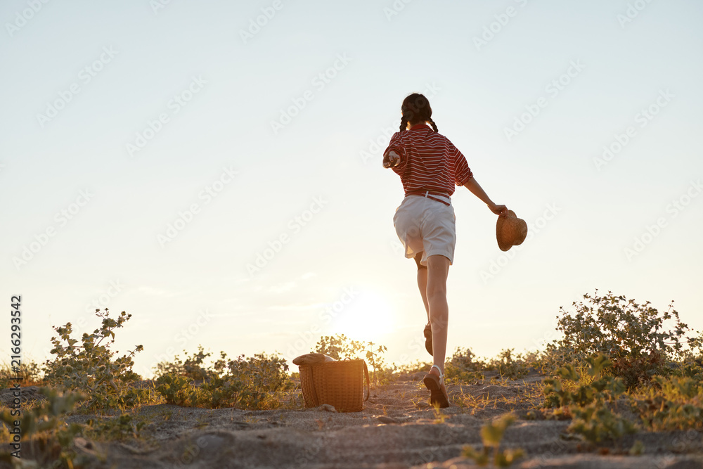 Fototapeta premium Young woman in a striped shirt and white shorts runs toward the setting sun on a dusty country trail, hat in hand, embodying carefree outdoor exploration.