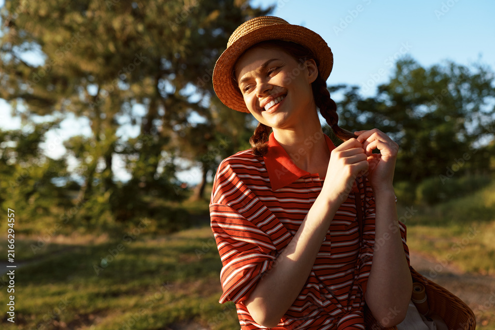 Naklejka premium Young woman in a striped red and white shirt smiles in bright sunlight in a park setting. Outdoor lifestyle portrait with natural light, casual mood, warm tones.