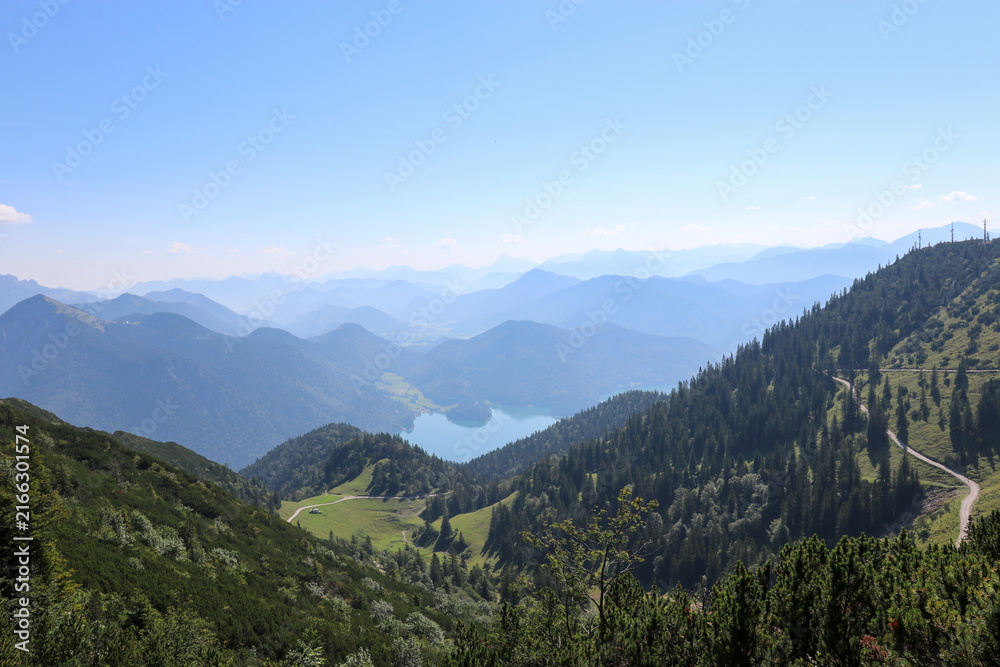 Fototapeta premium Atemberaubender Panoramablick auf einen Bergsee, eingebettet in ein grünes Tal der Alpen. Die blauen Bergschichten im Hintergrund unterstreichen die Weite und Schönheit dieser beeindruckenden Bergland