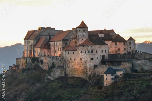 Historische Burganlage auf einem bewaldeten Hügel im warmen Abendlicht. Die alte Festung mit ihren Steinmauern und roten Dächern strahlt eine märchenhafte und erhabene Atmosphäre aus.