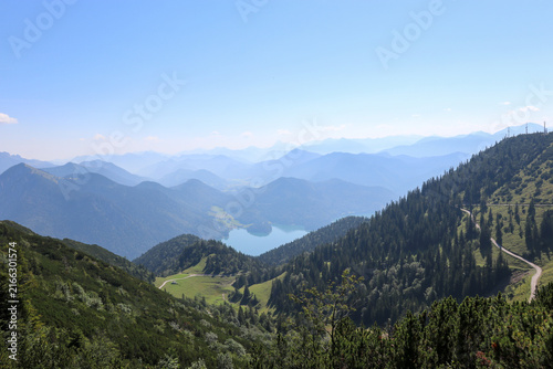 Atemberaubender Panoramablick auf einen Bergsee, eingebettet in ein grünes Tal der Alpen. Die blauen Bergschichten im Hintergrund unterstreichen die Weite und Schönheit dieser beeindruckenden Bergland